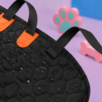 Close-up of a black quilted waterproof bag with orange accents and black webbing straps, shown on a purple surface with pink and blue toy props in the background.