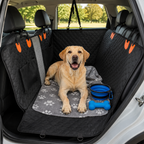 A yellow Labrador retriever lies in the back of a car trunk wearing a black quilted pet car seat cover with orange accents, placed over a gray paw-print mat. A blue collapsible bowl and blue bone toy are beside the dog.